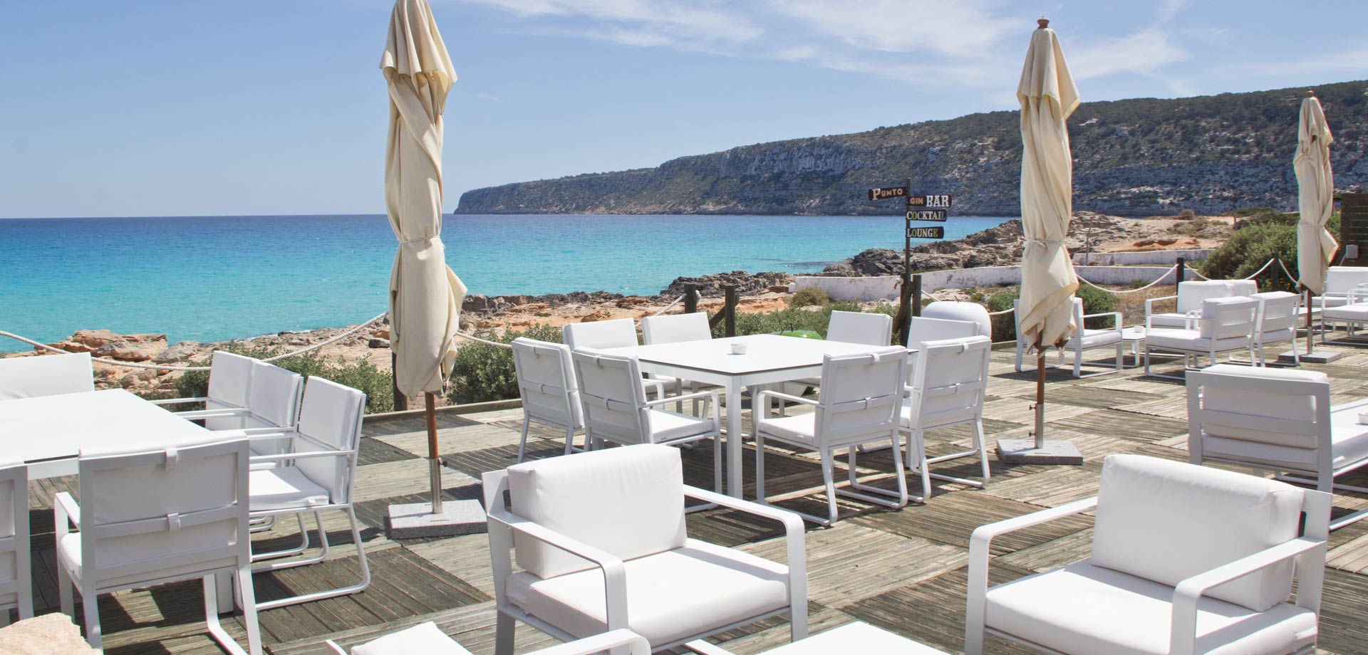 Restaurante Es Caló Formentera: verse vis aan het haventje van Sant Agustí 2 Witte loungestoelen en parasols op het terras van Restaurante Es Caló met de kliffen van La Mola en de zee