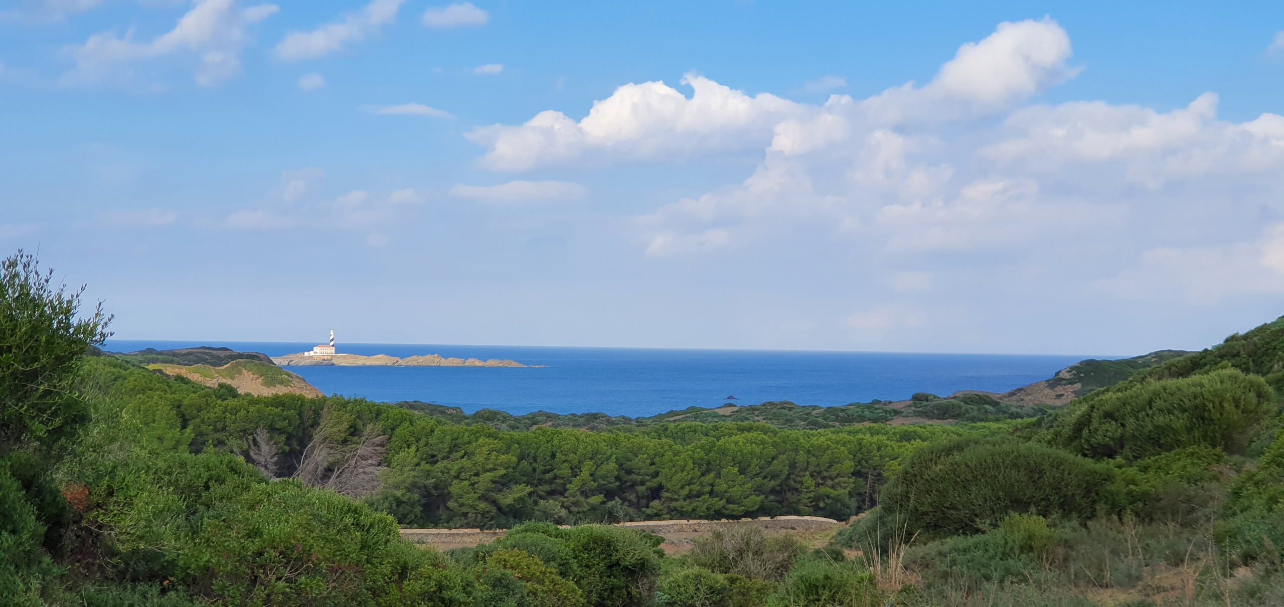 De baai van Es Grau op Menorca gezien vanaf het kustpad, met dennenbos op de voorgrond en het eilandje voor de kust