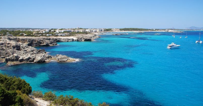De kust bij Es Pujols op Formentera met fel turquoise water, rotsranden en de lage witte bebouwing van het dorp erachter
