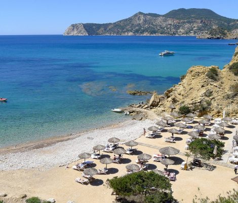 De kiezelbaai van Es Torrent op Ibiza van bovenaf, met strandparasols, helder water en de kliffen van Porroig