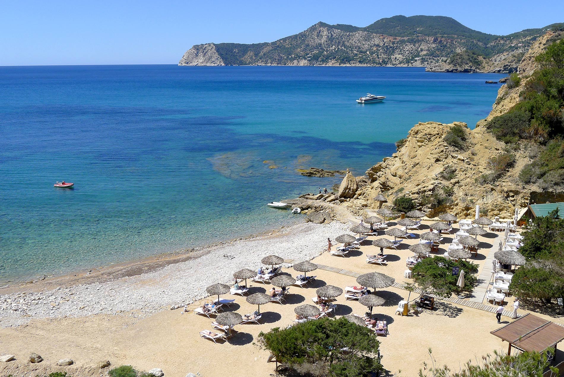 Es Torrent Ibiza: verborgen kiezelstrand in Porroig met topvis-restaurant 1 De kiezelbaai van Es Torrent op Ibiza van bovenaf, met strandparasols, helder water en de kliffen van Porroig