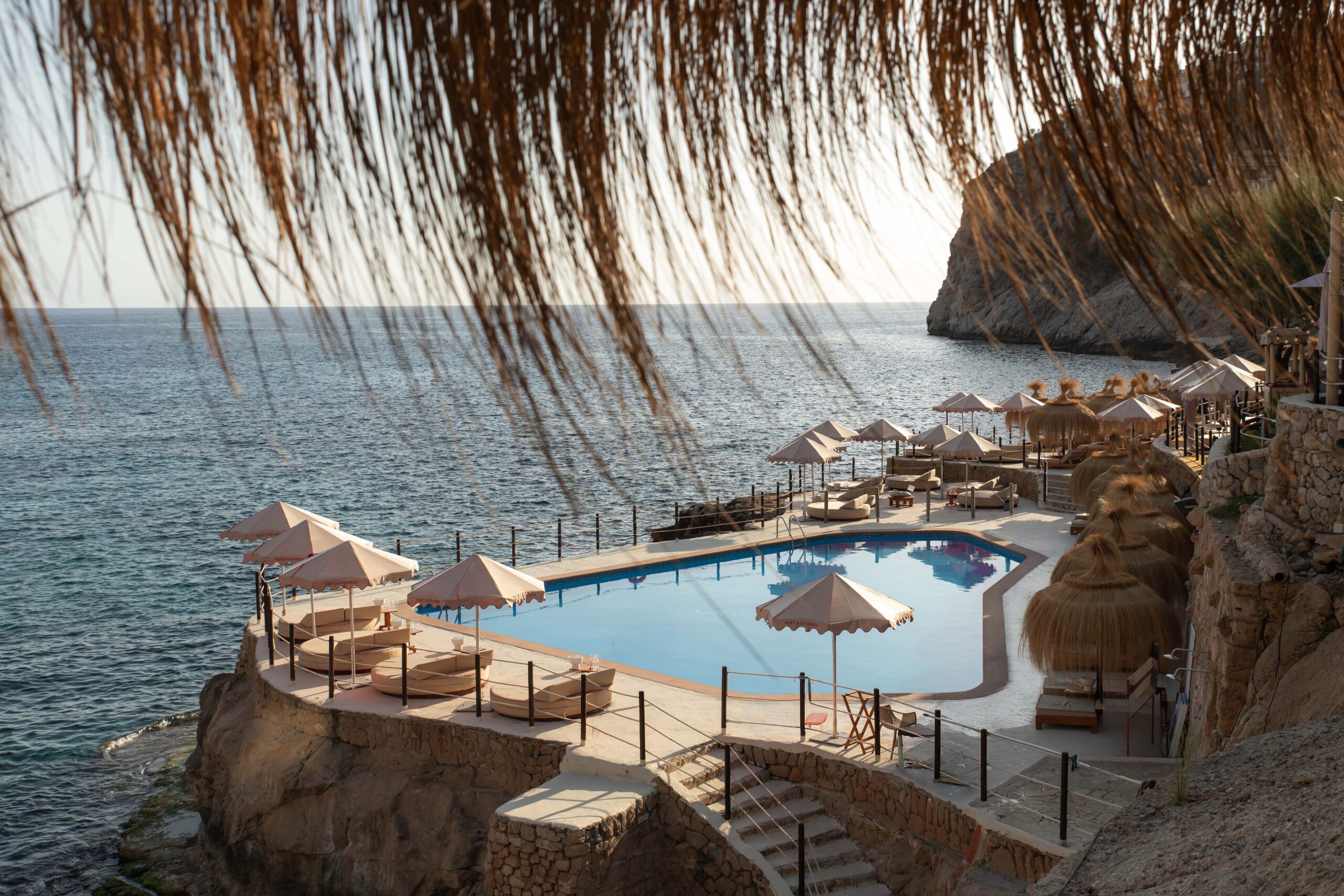 Gran Folies Mallorca: cliffside beachclub boven Cala Llamp bij Port d'Andratx 1 Het zwembad en de rieten parasols van Gran Folies op de klif boven Cala Llamp met de zee erachter