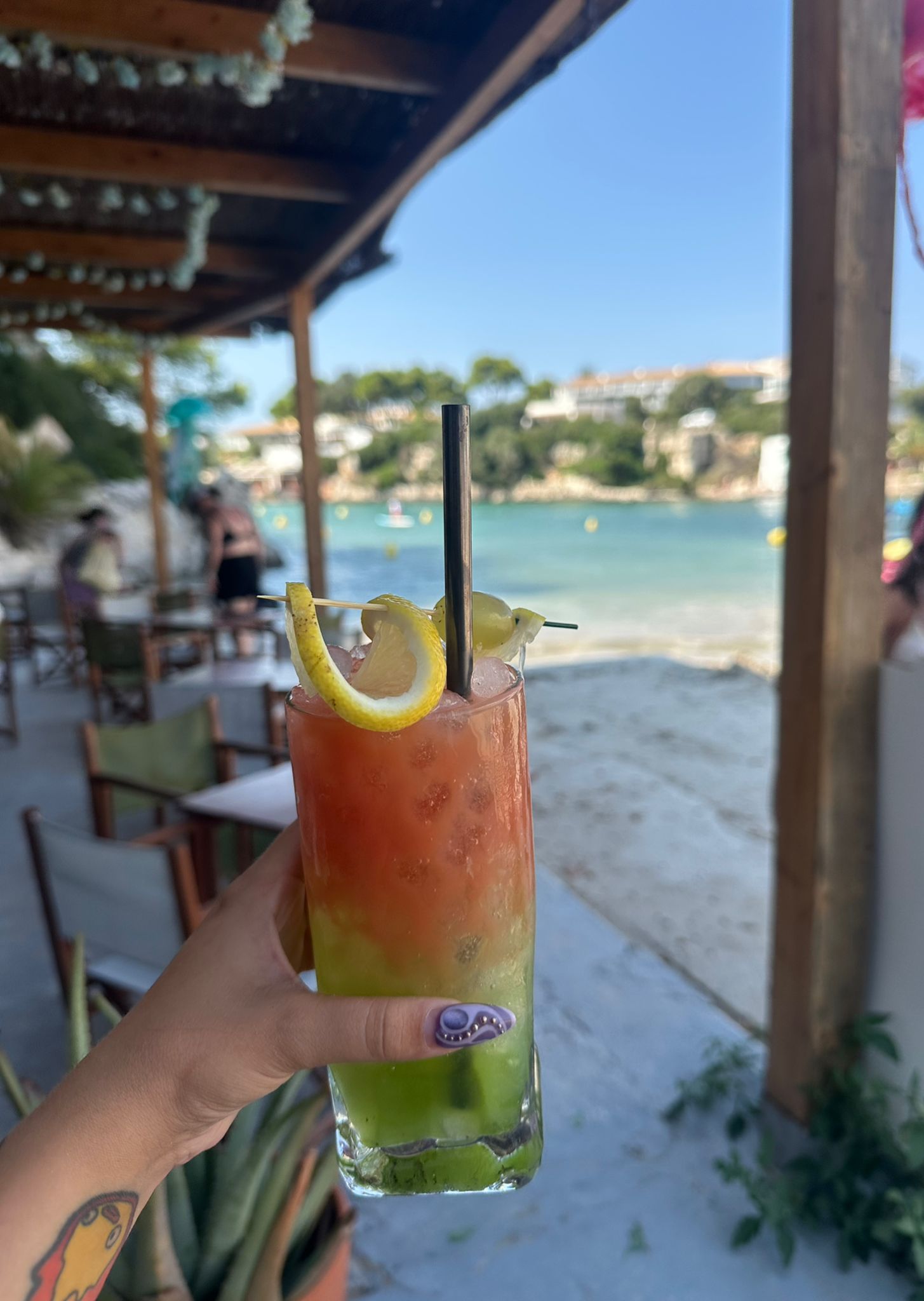 Kleurige cocktail in de hand bij Hopi Beach Bar met de cala op de achtergrond