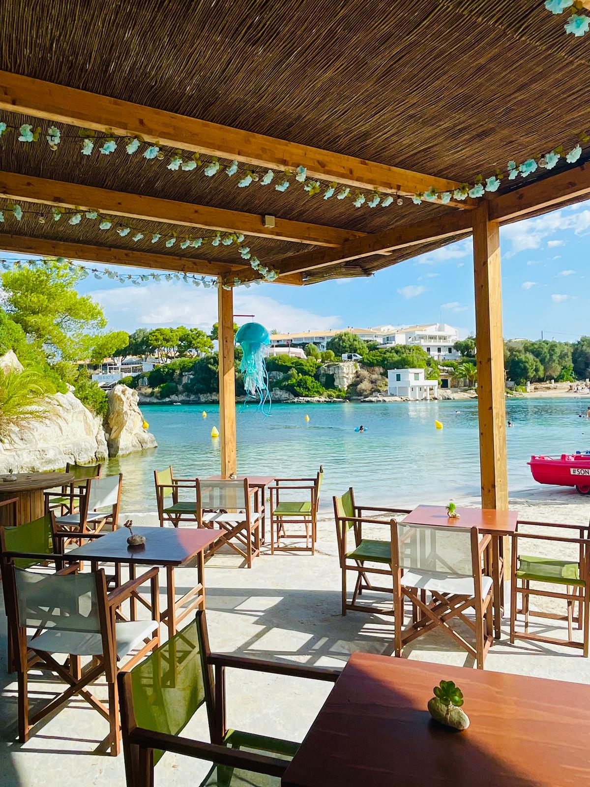 Het overdekte terras van Hopi Beach Bar met houten tafels en de kwal-decoratie boven de turquoise cala