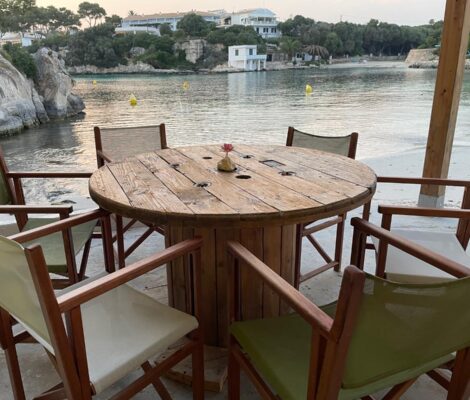 Houten strandtafel met groene regisseursstoelen op het terras van Hopi Beach Bar met de cala van Cala en Blanes erachter
