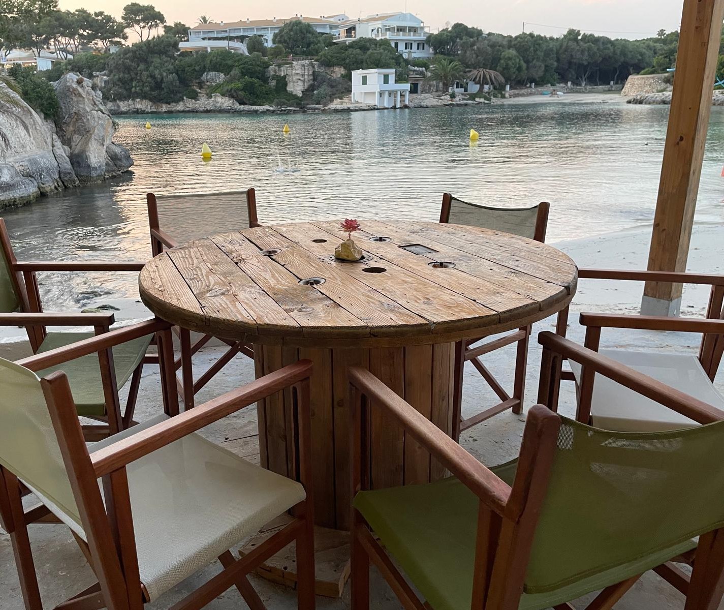 Houten strandtafel met groene regisseursstoelen op het terras van Hopi Beach Bar met de cala van Cala en Blanes erachter
