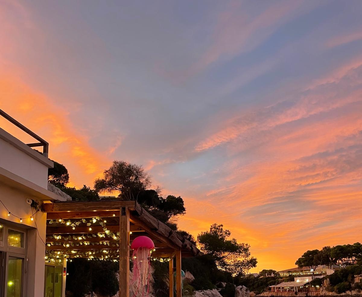 Het terras van Hopi Beach Bar met lichtjes en een roze kwal tegen een oranje avondlucht boven de cala