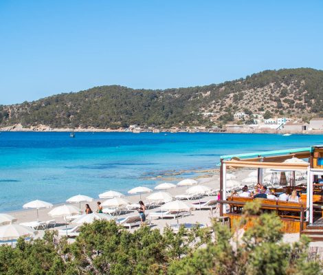 Het strandrestaurant Jockey Club aan het strand van Ses Salines, met witte parasols en de heuvels bij Sa Canal