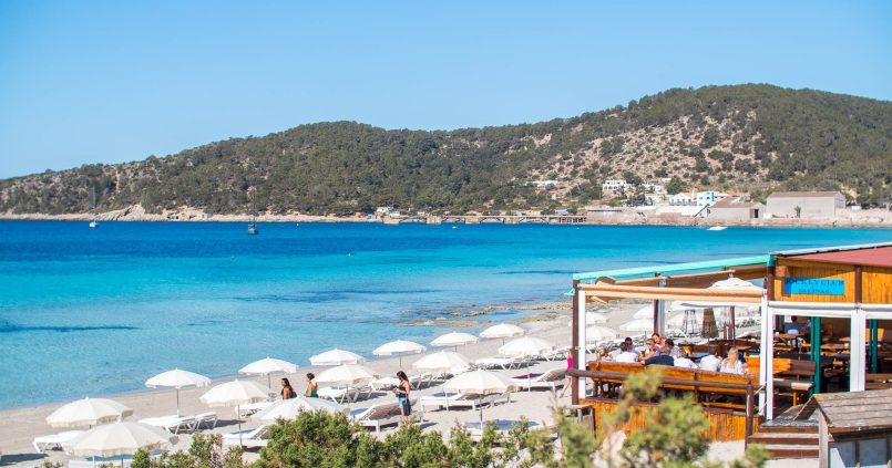 Het strandrestaurant Jockey Club aan het strand van Ses Salines, met witte parasols en de heuvels bij Sa Canal