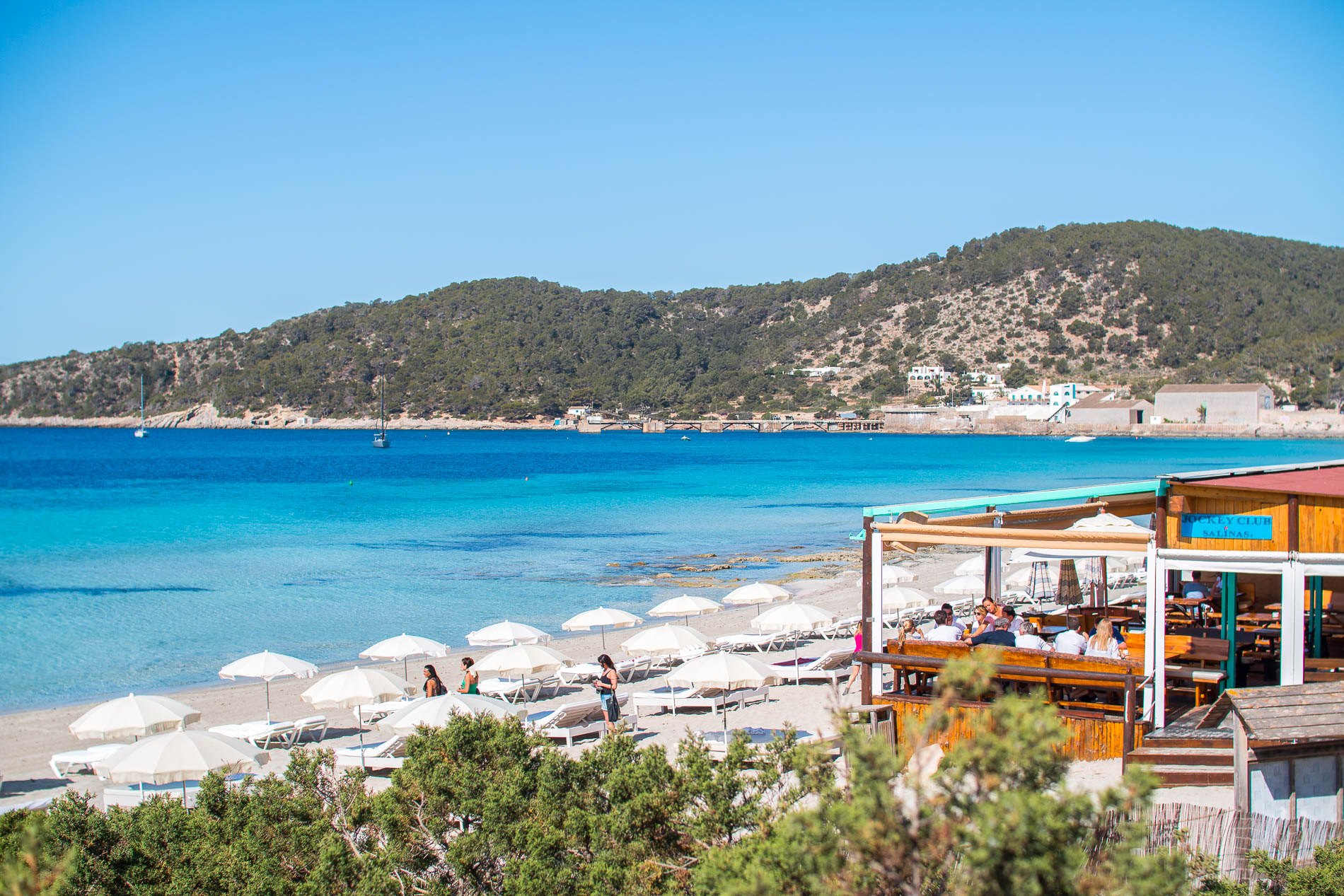 Het strandrestaurant Jockey Club aan het strand van Ses Salines, met witte parasols en de heuvels bij Sa Canal