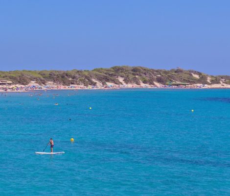 Het lange zandstrand van Ses Salines op Ibiza met turquoise water, zwemmers en de met pijnbomen begroeide duinen van het natuurpark