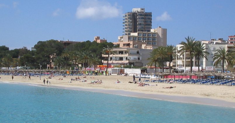 Het strand van Magaluf op Mallorca met turquoise water, rijen strohutparasols, palmen en de hotels van de badplaats erachter