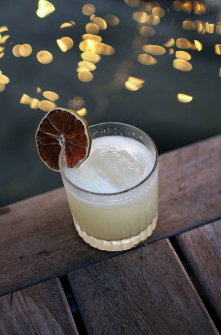 Een cocktail op een houten tafel bij Mariluz met warme avondlichtjes op de achtergrond