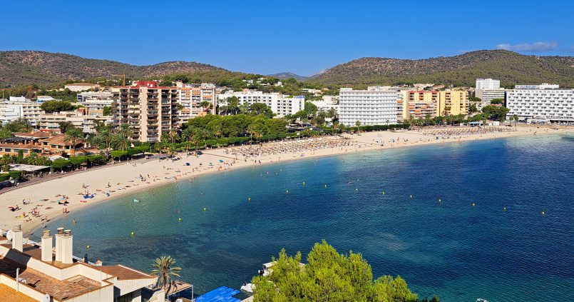 De baai van Palmanova op Mallorca met licht zand, helder blauw water, pijnbomen langs de promenade en hotels tegen de groene heuvels