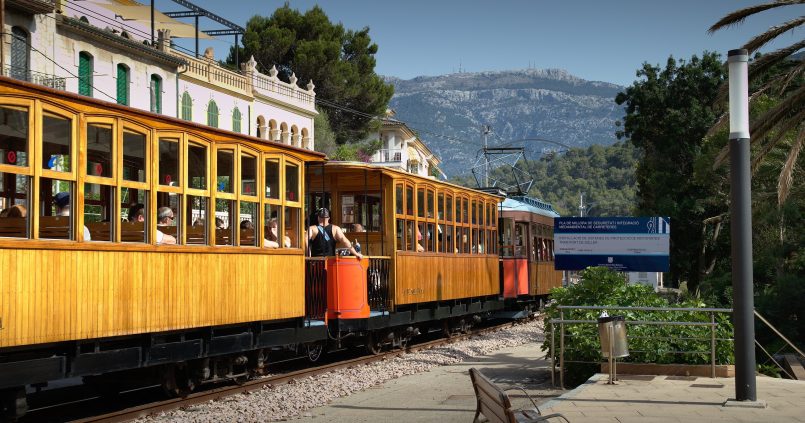 Houten tram met open rijtuigen vertrekt uit Port de Sóller richting Sóller, met de bergen van de Tramuntana op de achtergrond