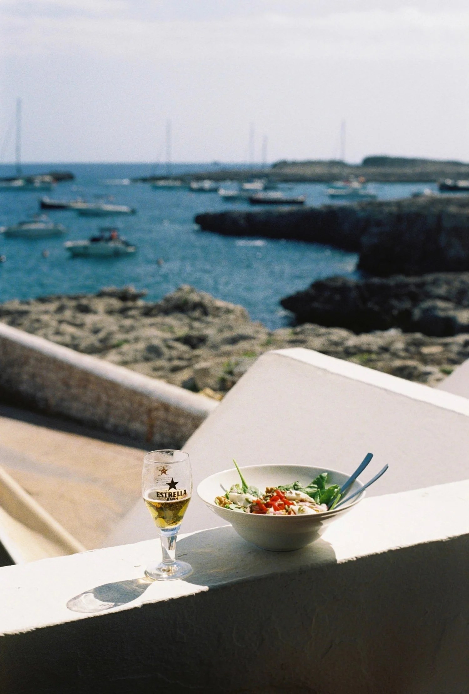 Een glas bier en een salade op het witte muurtje van Paupa met de baai en de bootjes van Cala Torret erachter