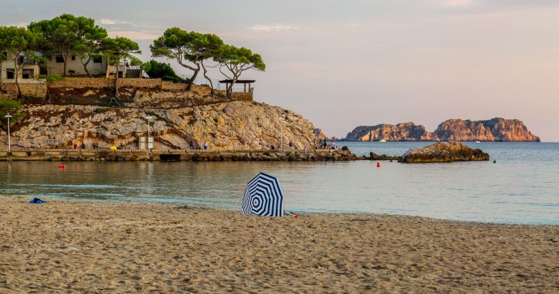 Het strand van Peguera op Mallorca in het avondlicht, met een gestreepte parasol op het zand, de rotspunt met parasoldennen en de eilandjes voor de kust