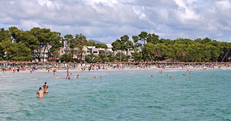 Badgasten in het ondiepe turquoise water van Platja d'Alcúdia op Mallorca, met pijnbomen en hotels achter het strand