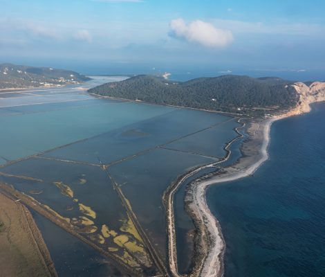 Luchtfoto van Platja des Codolar op Ibiza: een lange kiezelstrook tussen de zoutpannen van Ses Salines en de zee, met de kliffen van Cap des Falcó