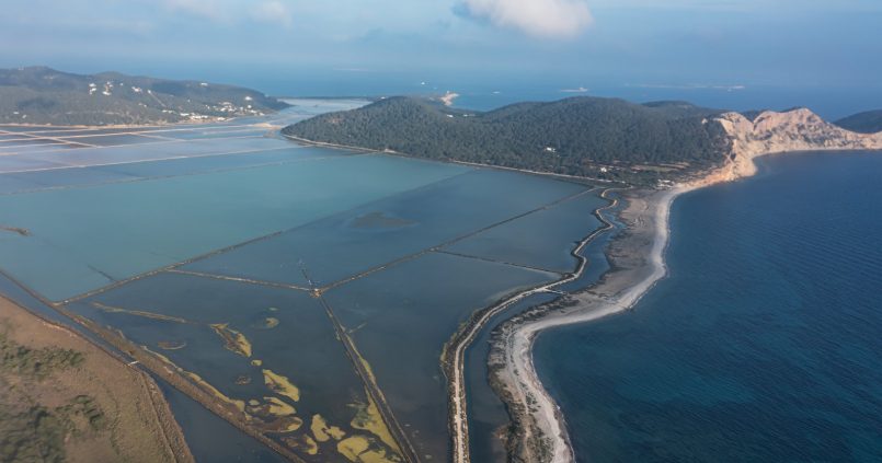 Luchtfoto van Platja des Codolar op Ibiza: een lange kiezelstrook tussen de zoutpannen van Ses Salines en de zee, met de kliffen van Cap des Falcó