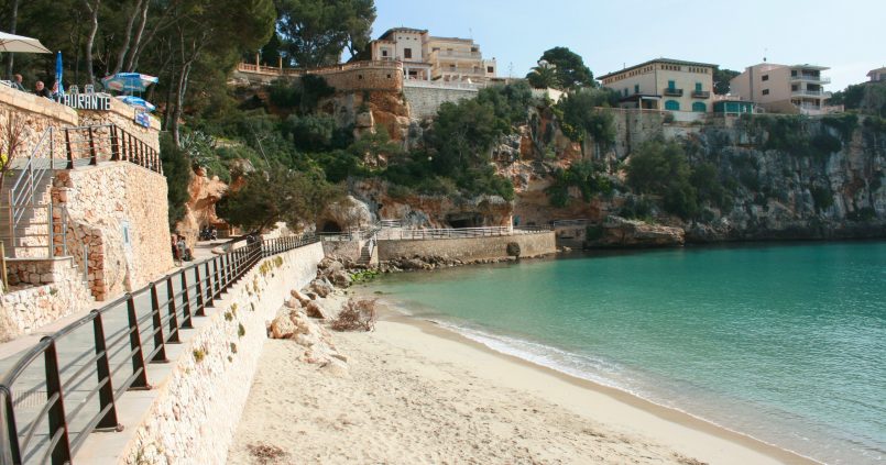 Het strand van Porto Cristo op Mallorca met turquoise water in een smalle inham, een stenen promenade en kliffen met huizen erboven
