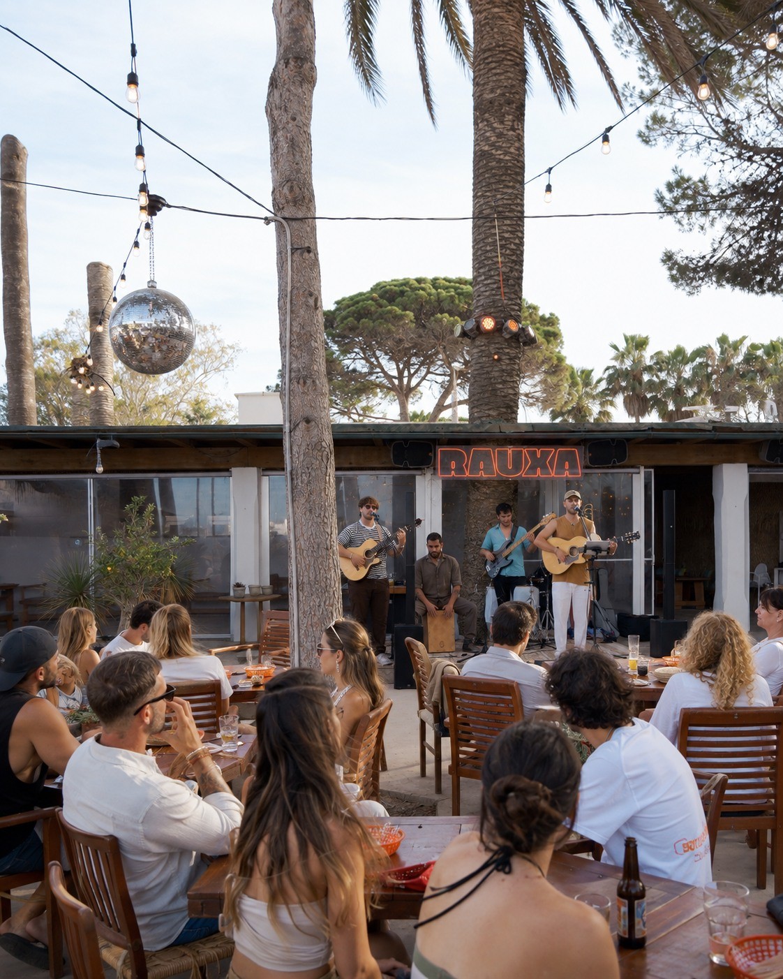 Het terras van Rauxa Ibiza met houten tafels, palmen en een discobal in de avond