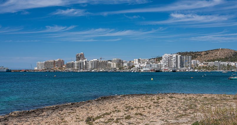 De baai van Sant Antoni op Ibiza met de skyline van de stad, helder blauw water en rotsen op de voorgrond