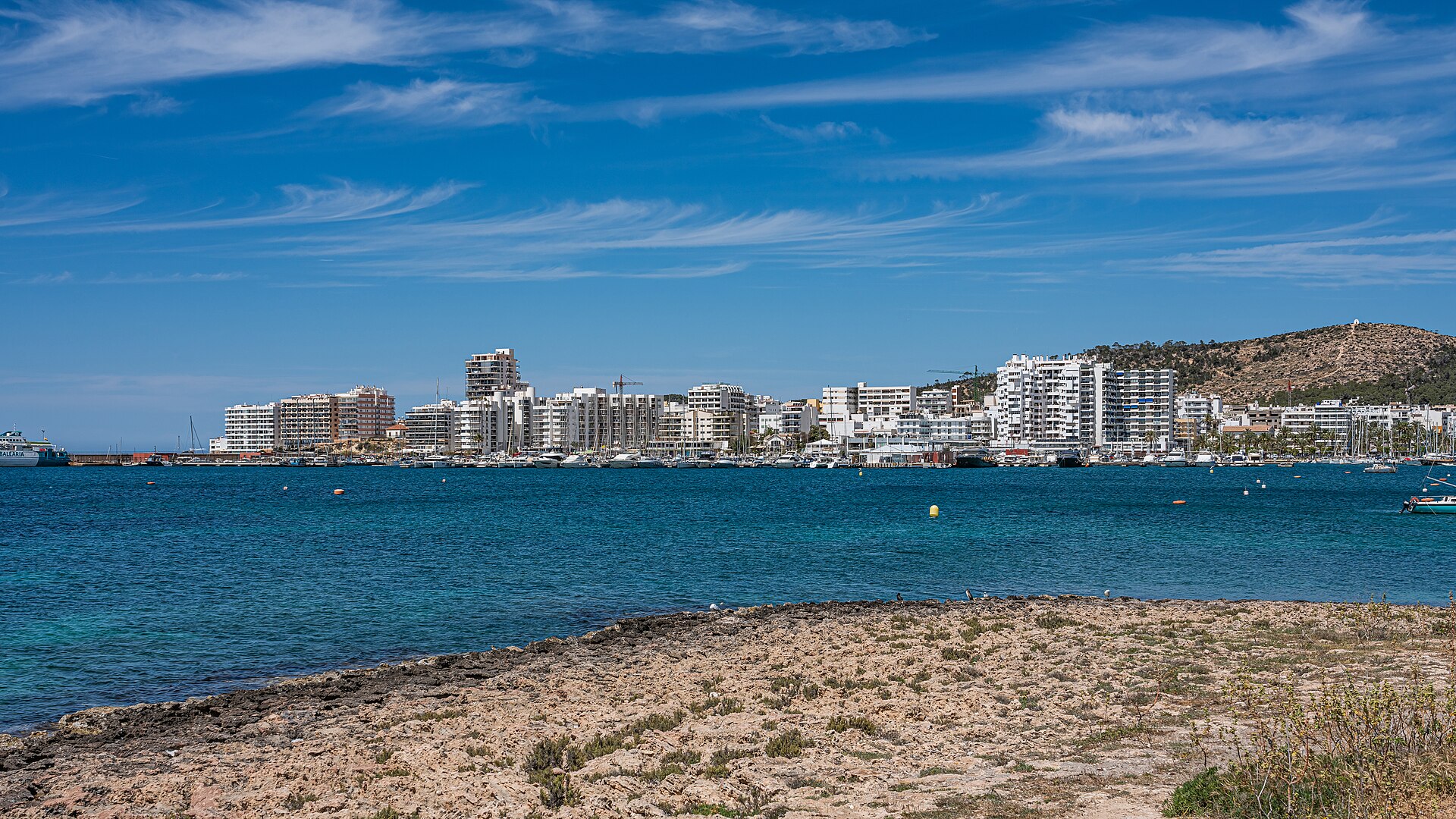 De baai van Sant Antoni op Ibiza met de skyline van de stad, helder blauw water en rotsen op de voorgrond