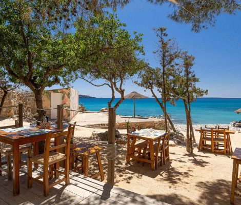 Houten tafels onder pijnbomen op het terras van Ses Boques, met het kiezelstrand, parasols en turquoise zee erachter