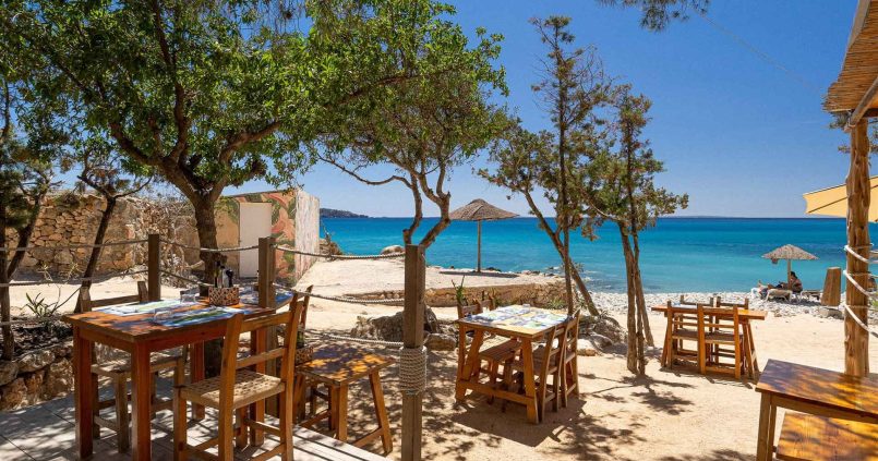 Houten tafels onder pijnbomen op het terras van Ses Boques, met het kiezelstrand, parasols en turquoise zee erachter