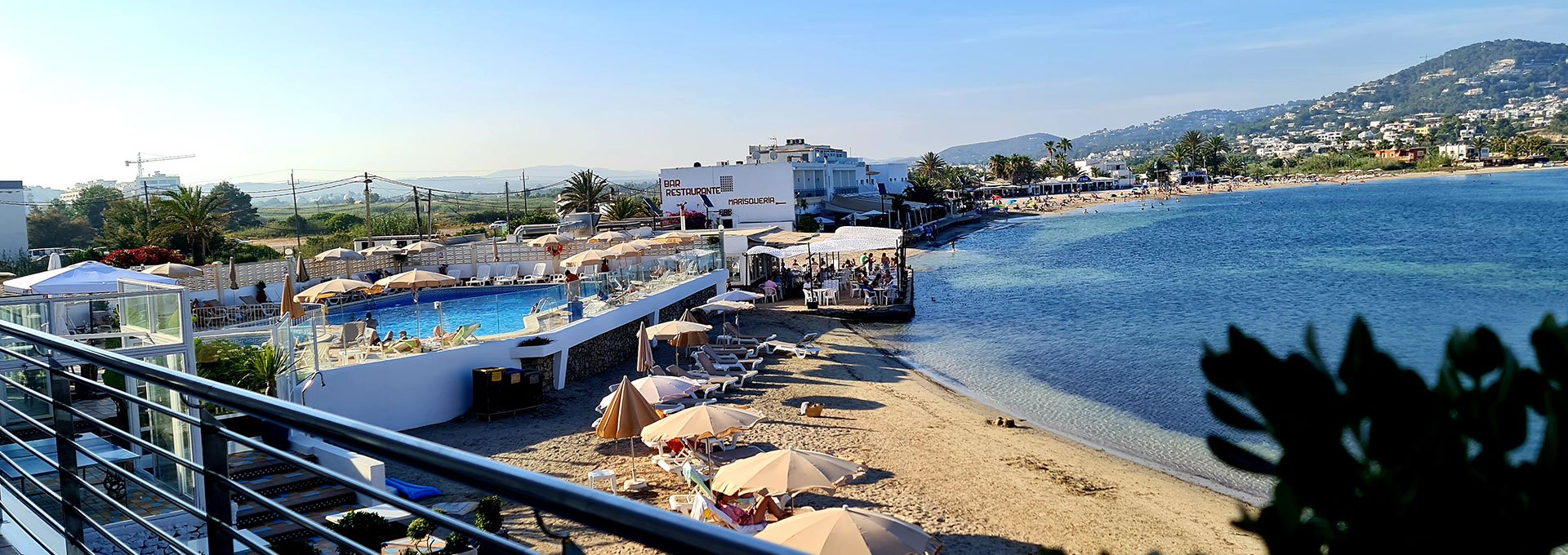 Het zandstrand van Talamanca met parasols, ondiep helder water en de heuvels aan de oostkant van de baai, gezien vanaf Hotel Argos