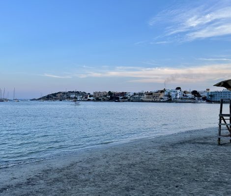 Het zandstrand van Talamanca bij Ibiza-stad in het avondlicht, met een reddingspost, kalm water en de huizen rond Marina Botafoch in de verte