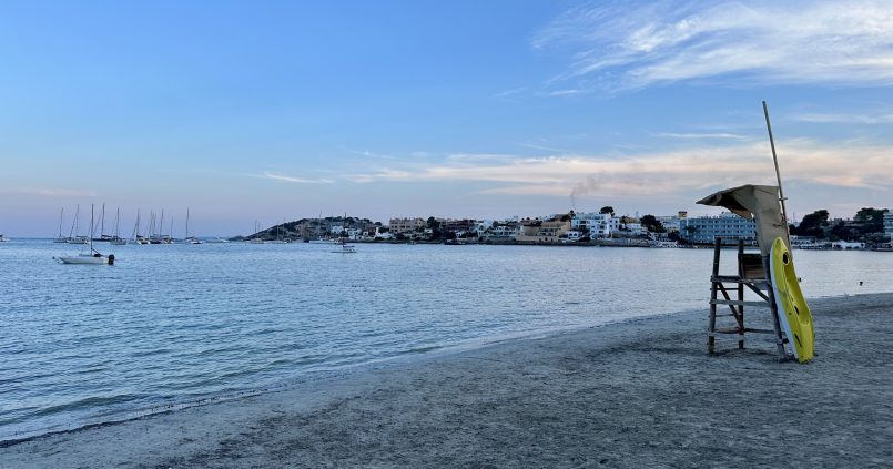 Het zandstrand van Talamanca bij Ibiza-stad in het avondlicht, met een reddingspost, kalm water en de huizen rond Marina Botafoch in de verte