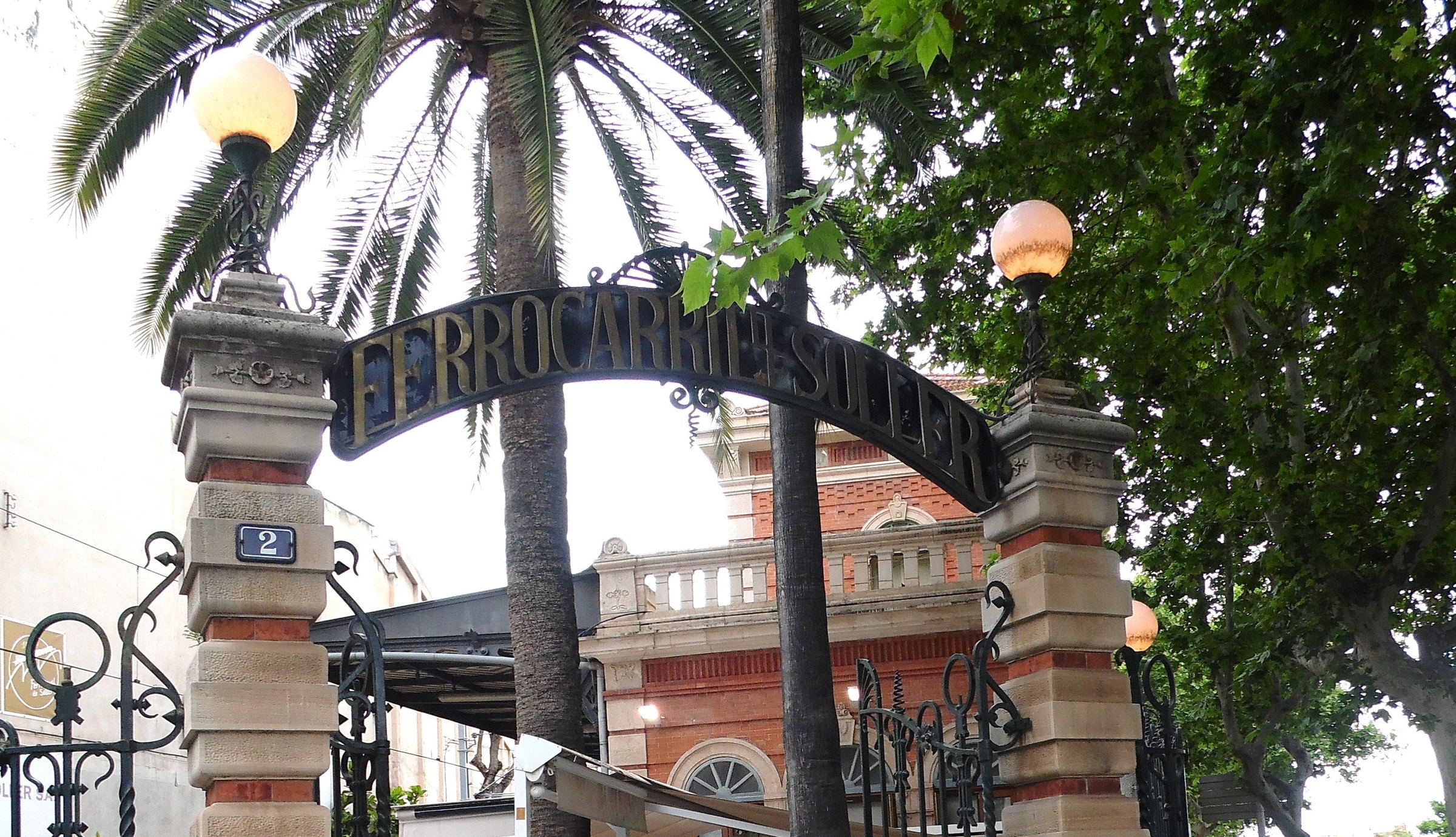 Trein Palma Sóller: het houten treintje door de bergen naar Sóller en de haven 2 De smeedijzeren toegangspoort met de letters Ferrocarril de Sóller bij het station in Palma, met een palm en het bakstenen stationsgebouw erachter
