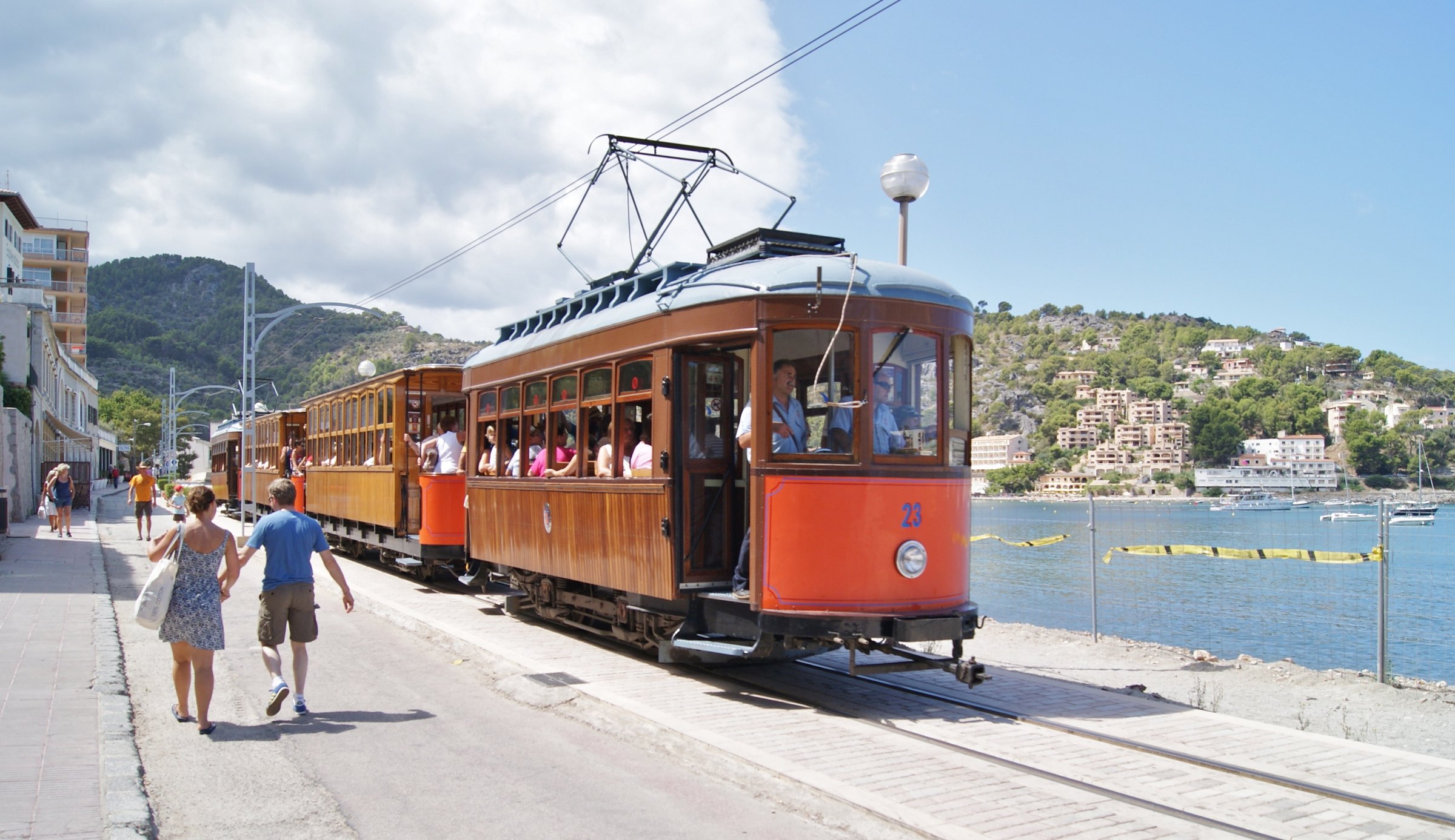 Trein Palma Sóller: het houten treintje door de bergen naar Sóller en de haven 4 De houten tram van Sóller met oranje front rijdt langs de baai van Port de Sóller, met beboste bergen op de achtergrond