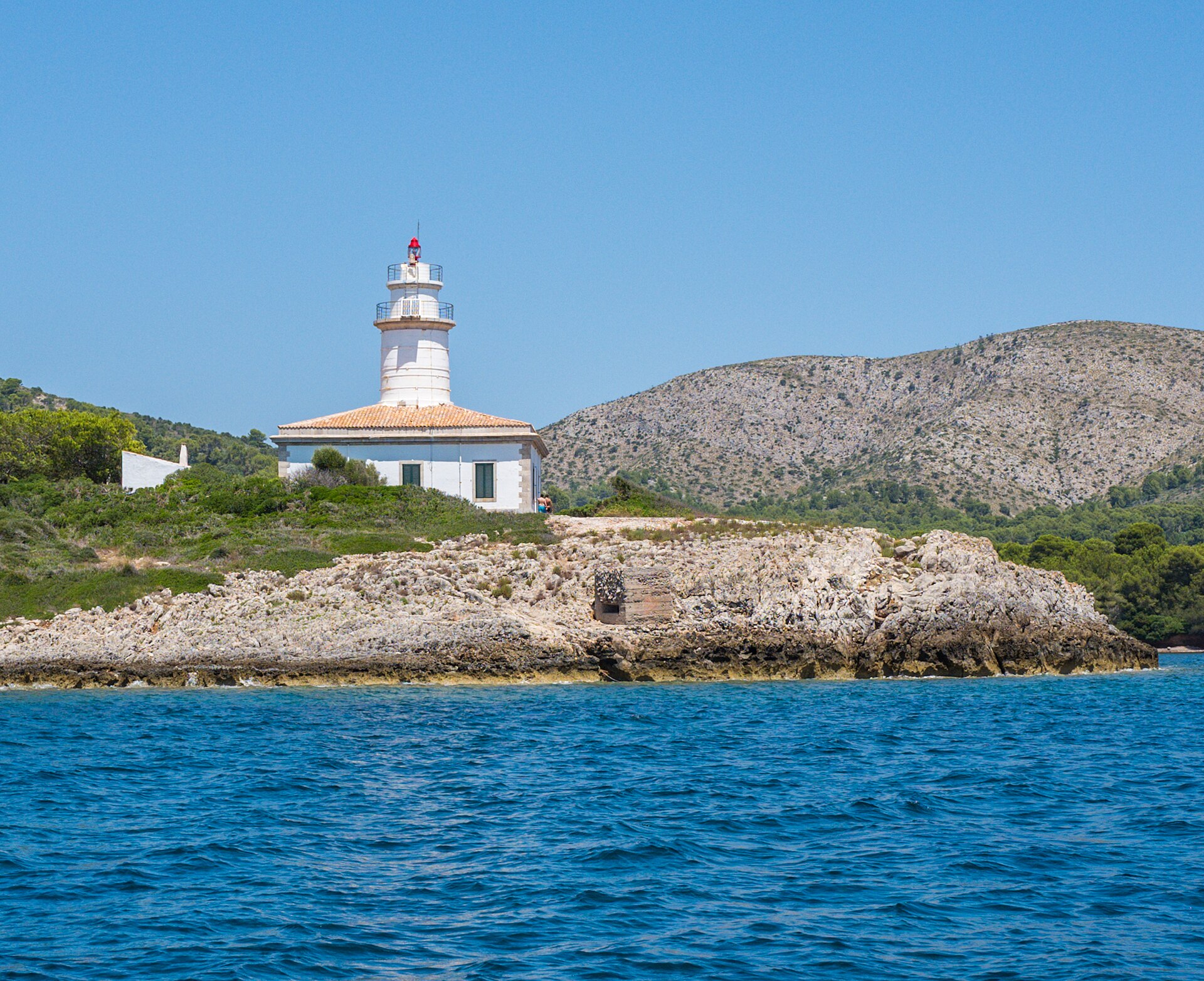 Alcanada Mallorca: rustig snorkelstrand met vuurtoreneilandje 5 De witte vuurtoren met het okerkleurige dak van het wachtershuis op de rotsige kust van Illa d'Alcanada, gezien vanaf het water