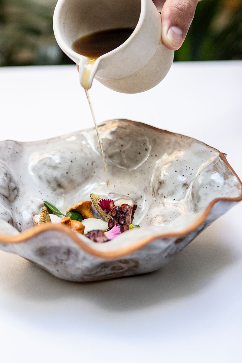 Een bouillon wordt uit een aardewerken kannetje geschonken over een gerecht met zeevruchten en bloemen in een golvende schaal bij Aromata in Palma