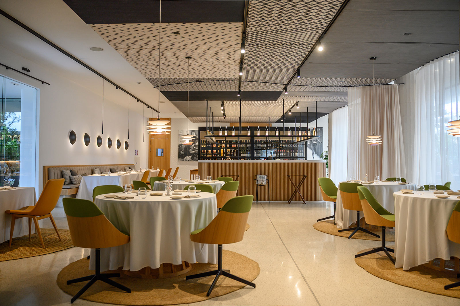 De eetzaal van restaurant Aromata in Palma met ronde tafels, groene en oranje stoelen en op de achtergrond de bar met wijnrekken