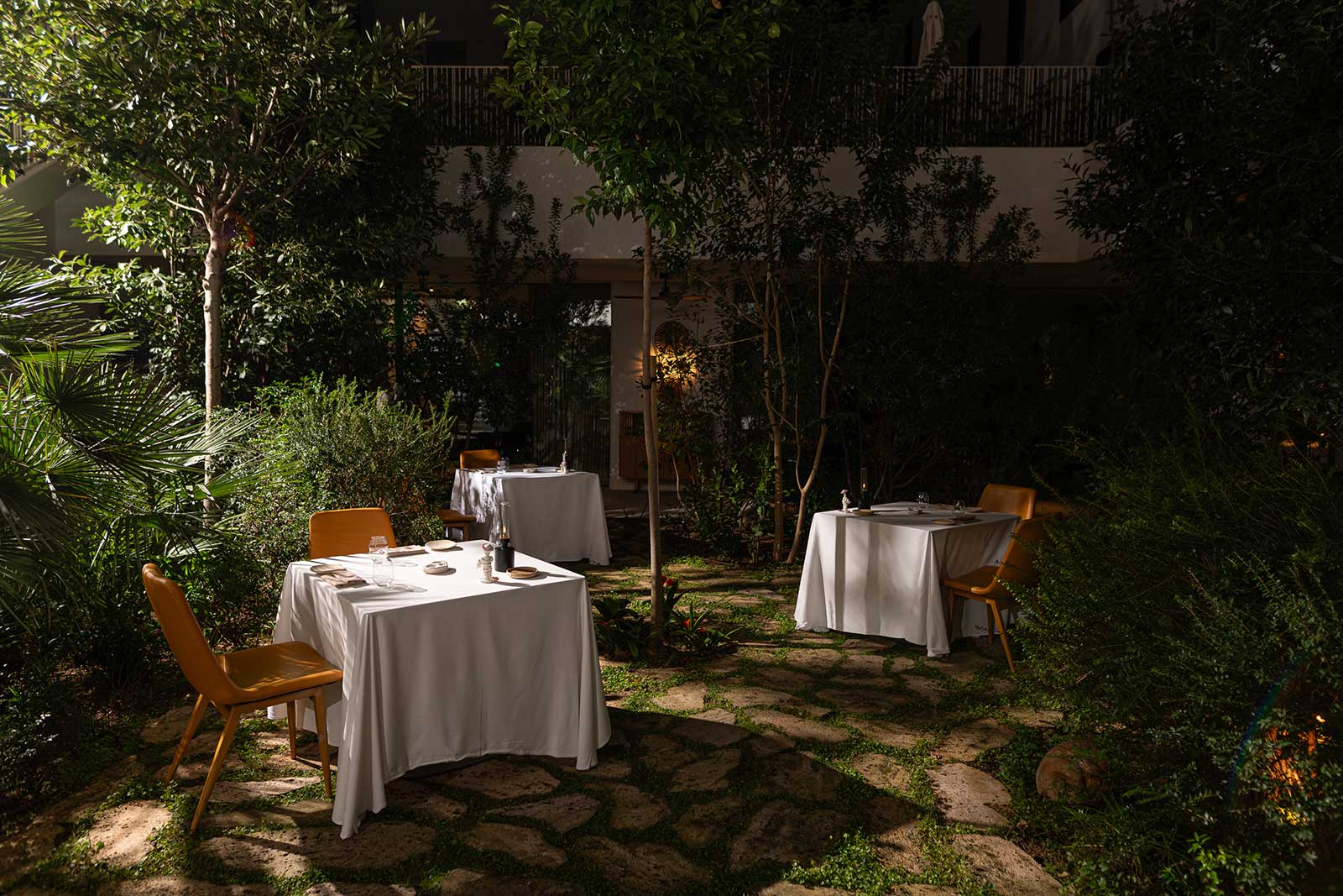 Gedekte tafels met witte kleden in de verlichte tuin van restaurant Aromata in Palma, tussen bomen en struiken in de avond