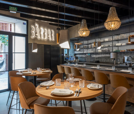De eetzaal van restaurant BàrBar in Palma met het neonlogo aan de balk, ronde houten tafels, bruine stoelen en de lange keukenbar met rieten hanglampen
