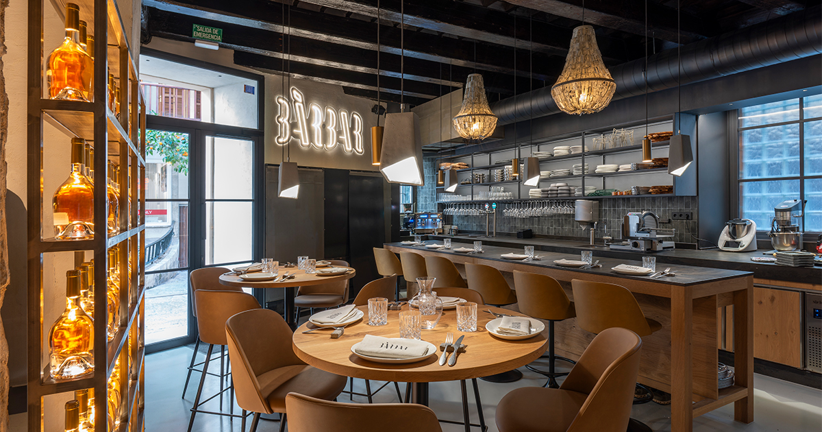 De eetzaal van restaurant BàrBar in Palma met het neonlogo aan de balk, ronde houten tafels, bruine stoelen en de lange keukenbar met rieten hanglampen