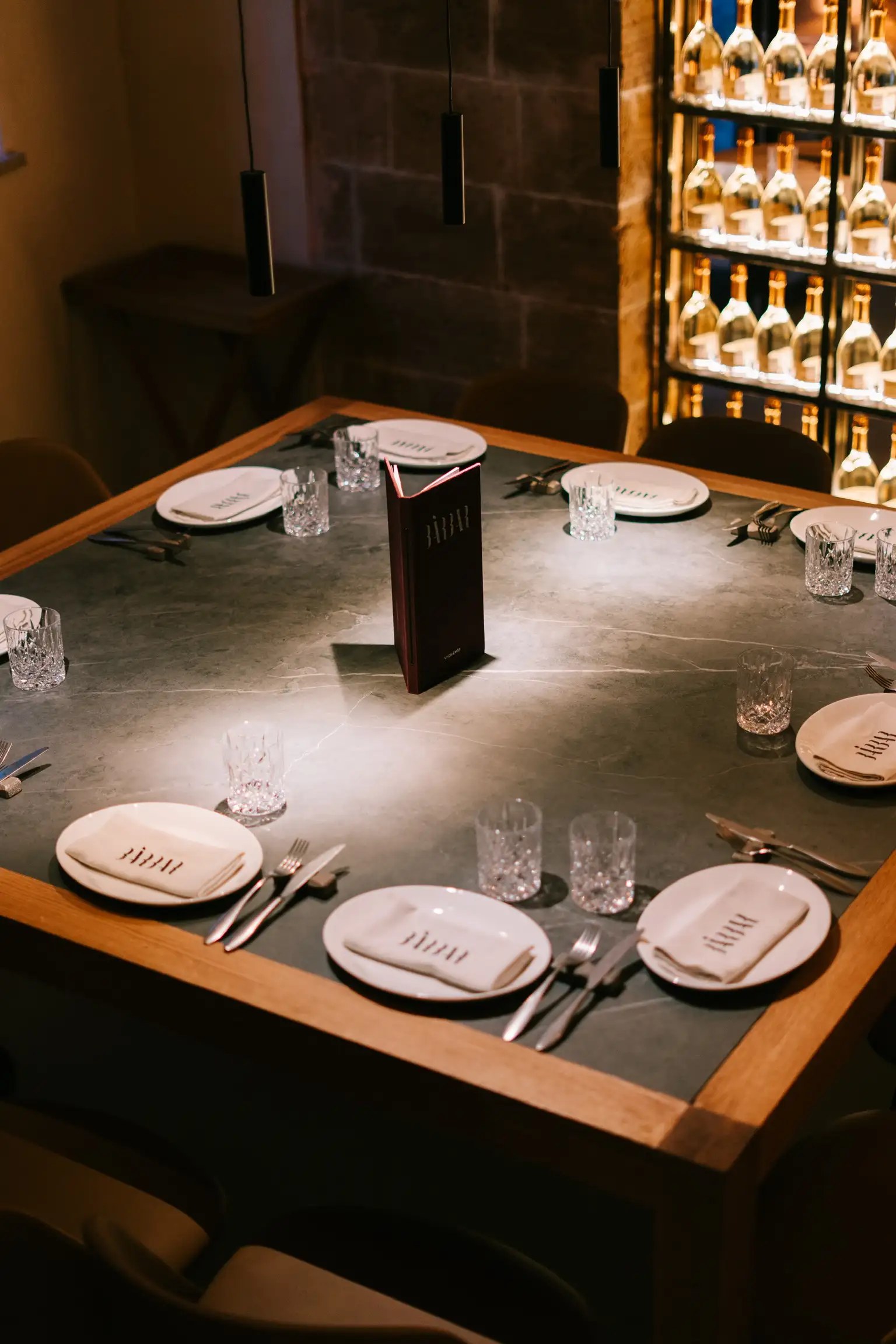 Vierkante tafel bij BàrBar in Palma, gedekt voor zes personen met servetten met het logo, naast de verlichte wand met wijnflessen