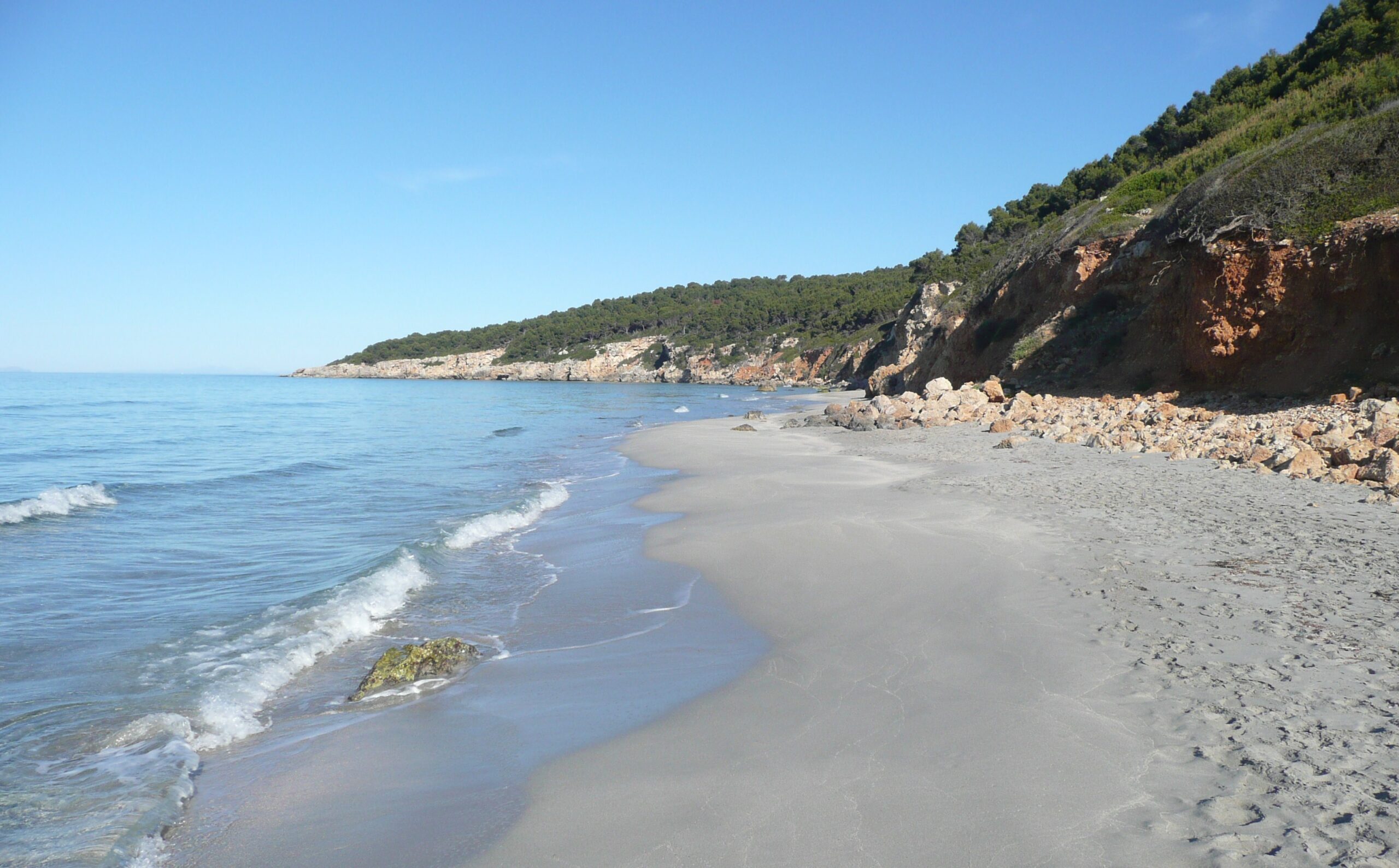 Binigaus Menorca: het wilde strand, het ravijn en de Cova des Coloms 1 Het lege zandstrand van Binigaus op Menorca met de rode rotsrand en pijnbomen erboven