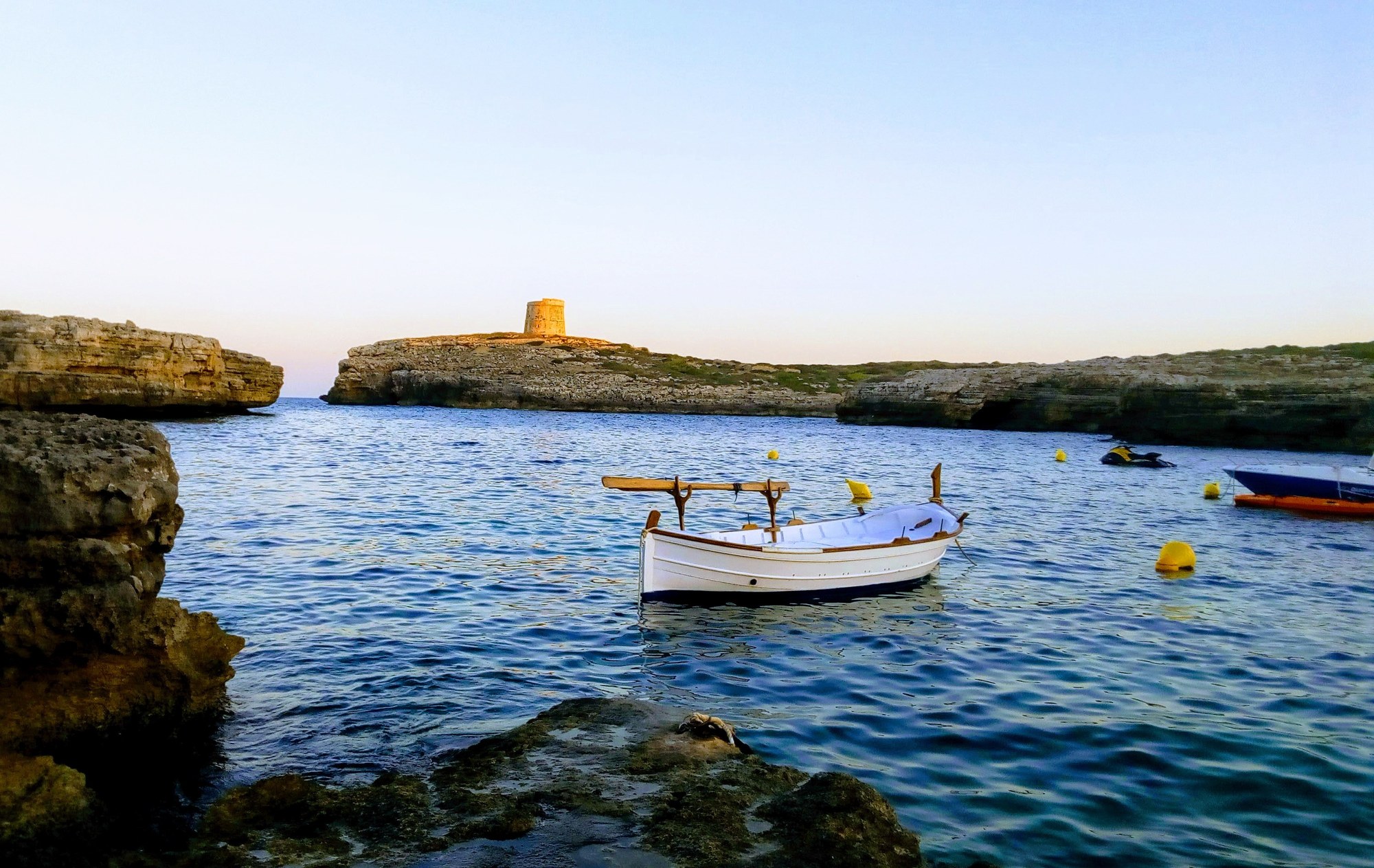 Witte llaüt voor anker in Cala Alcalfar met de Britse wachttoren op de kaap erachter, bij zonsondergang