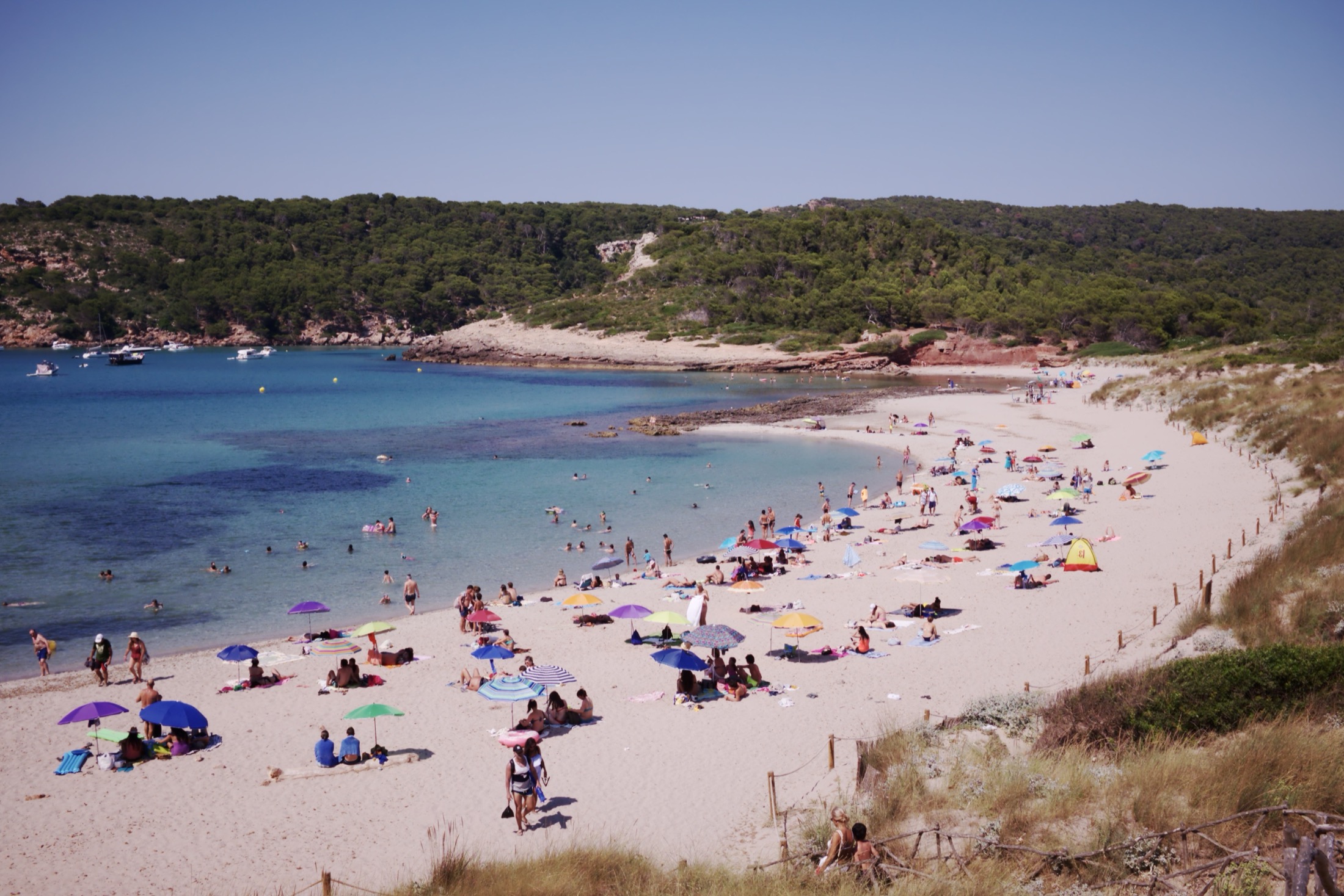 Het strand van Cala Algaiarens op een zomerdag, gezien vanaf het duinpad met parasols op het zand