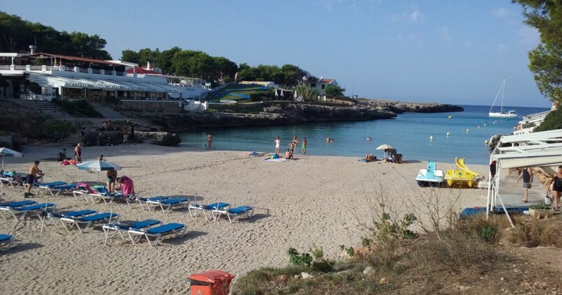 Cala Blanca Menorca: witte zandbaai met zonsondergang bij Ciutadella 1 Het witte zandstrand van Cala Blanca op Menorca met blauwe ligbedden, waterfietsen en de monding van de baai naar open zee