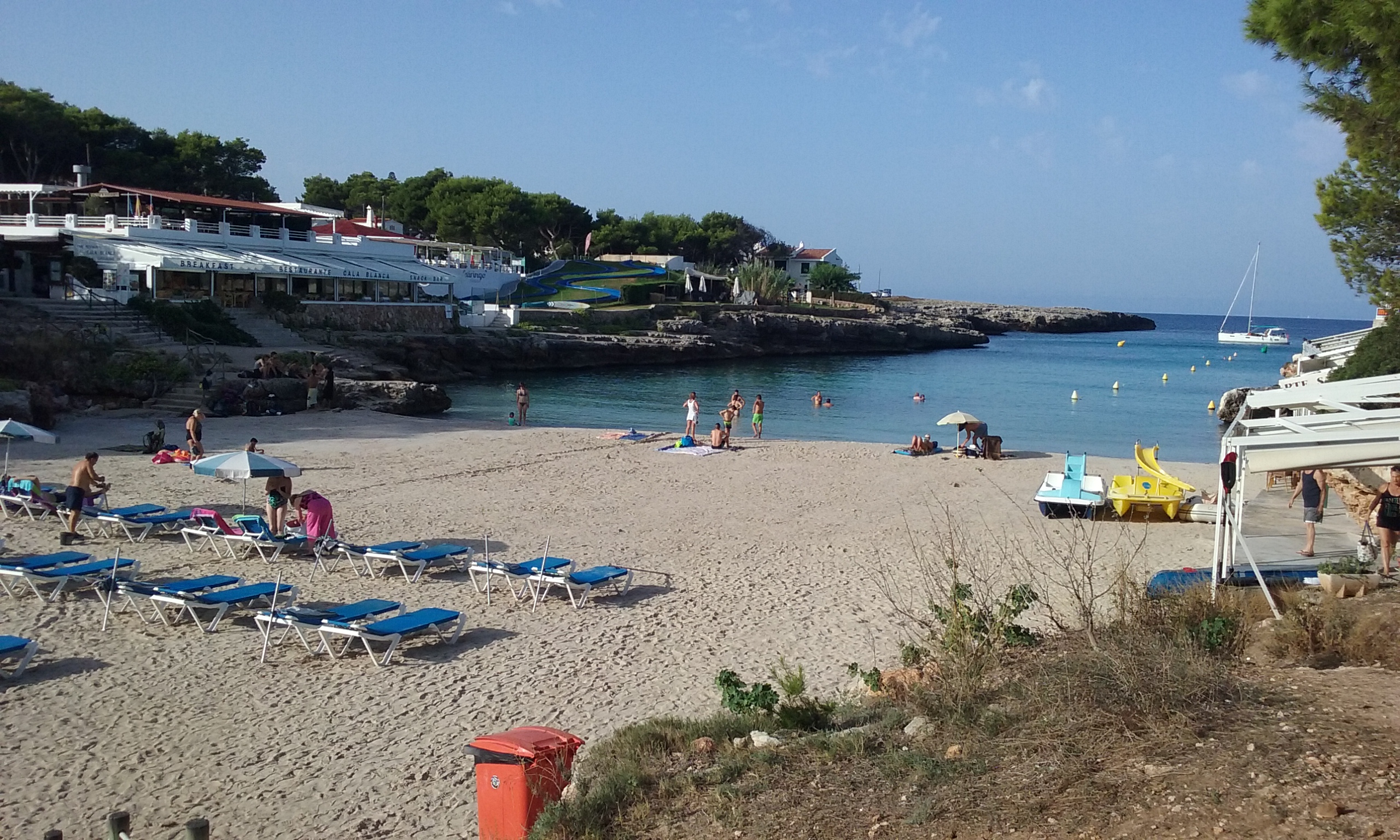 Cala Blanca Menorca: witte zandbaai met zonsondergang bij Ciutadella 1 Het witte zandstrand van Cala Blanca op Menorca met blauwe ligbedden, waterfietsen en de monding van de baai naar open zee