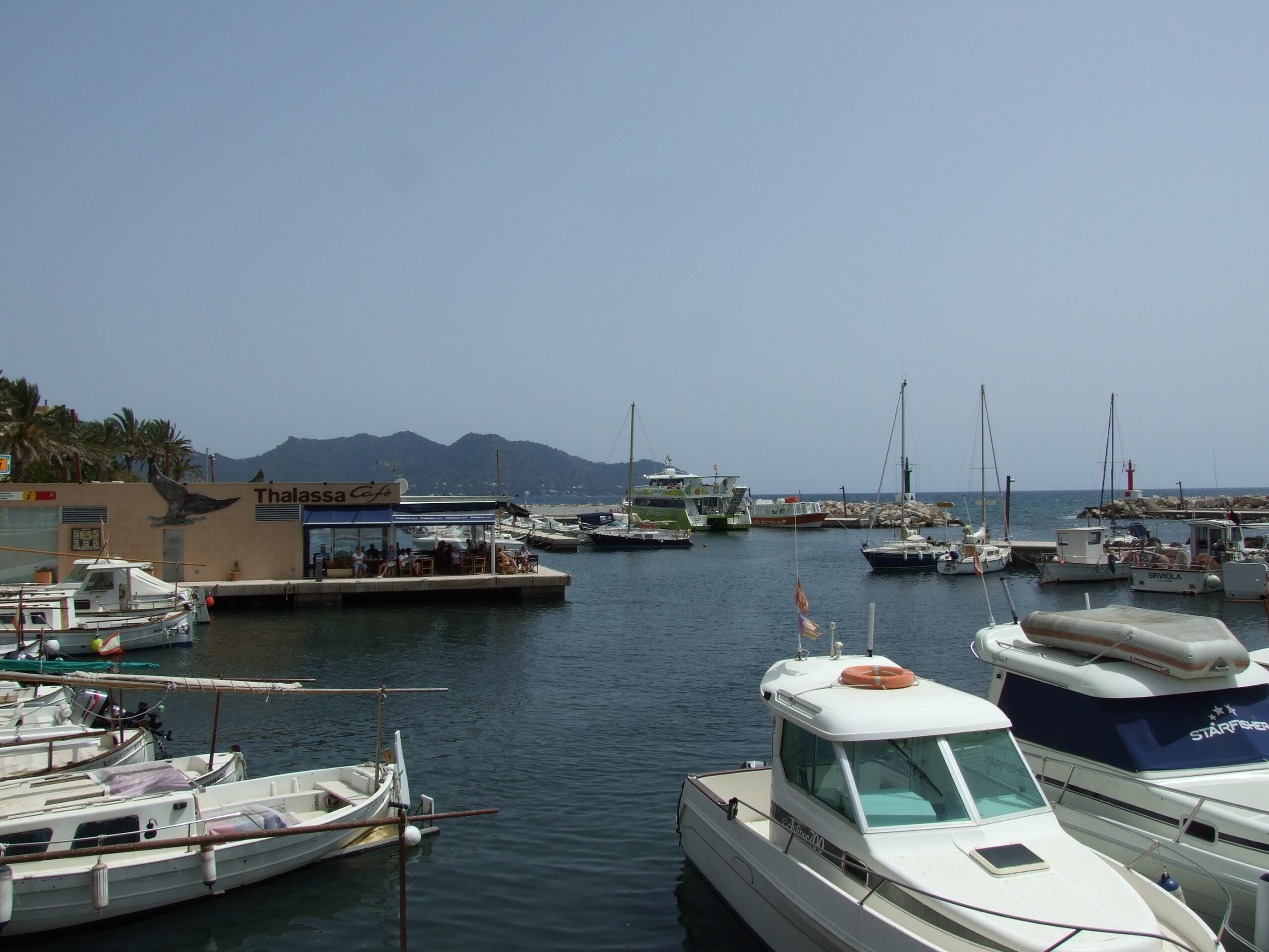De havenkom van Cala Bona met kleine open houten vissersboten op de voorgrond, plezierjachten erachter en de havenmond met de bergen van de Serra de Llevant in de verte