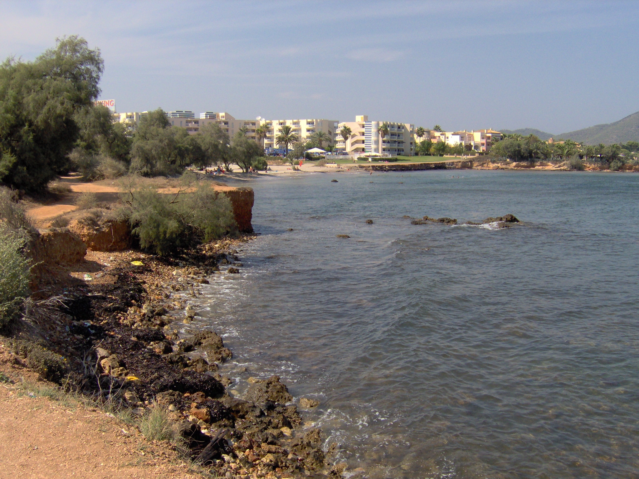 De roodbruine rotsoever tussen Cala Bona en Port Nou, met een klein strandje en de lage bebouwing van de badplaats erachter