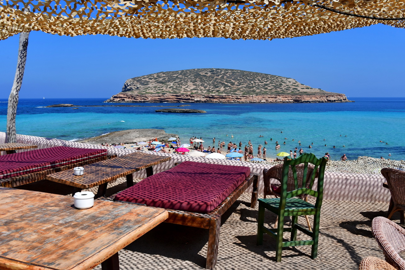 Uitzicht vanonder het rieten zonnedak van de strandtent op Cala Comte, met het drukke zandstrandje en s'Illa des Bosc achter het turquoise ondiepe water